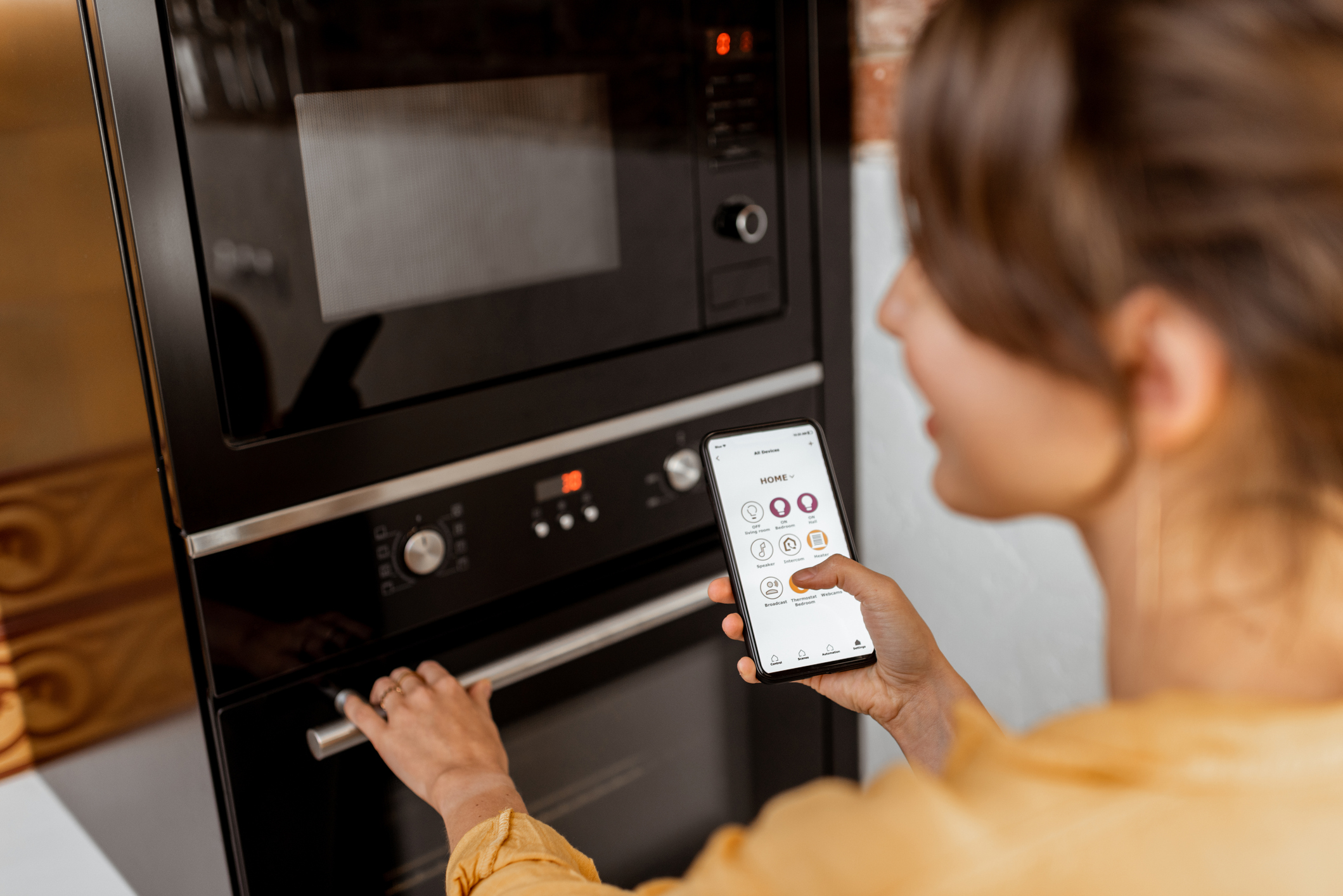 Homeowner using a smartphone to control a built-in kitchen oven, showing smart kitchen technology integration for convenience and efficiency.