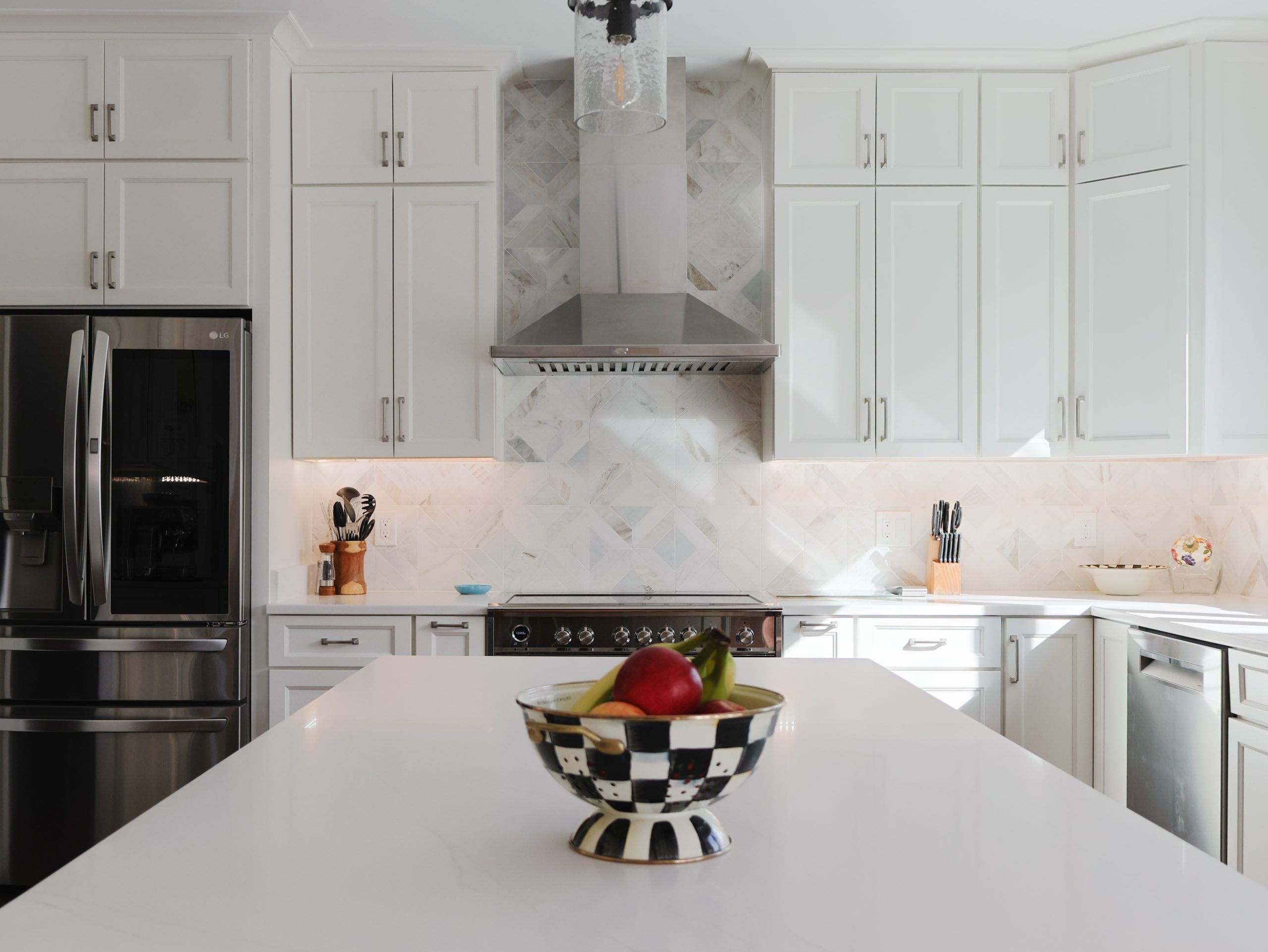 Bright kitchen with white wall-mounted cabinets, stainless steel range hood, marble tile backsplash, and island with fruit bowl in the foreground.