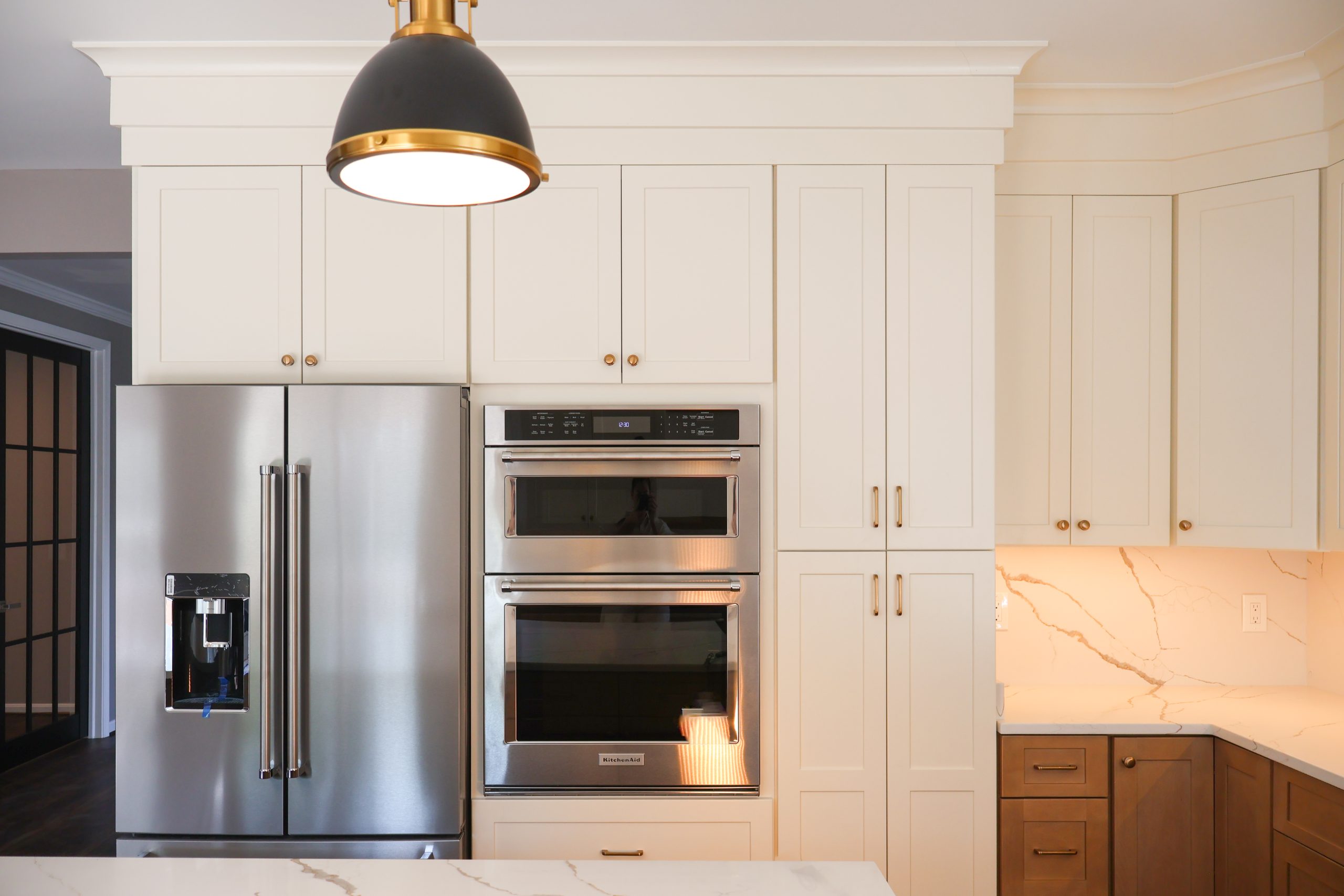 White wall-mounted kitchen cabinets with brass handles, built-in double oven, stainless steel refrigerator, and marble backsplash under warm lighting.